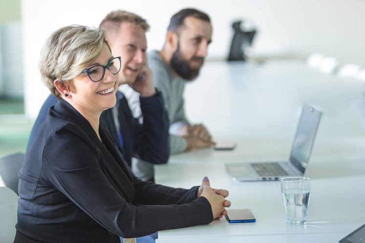 A woman and two men sitting at a conference table with a laptop and a glass of water in front of them.