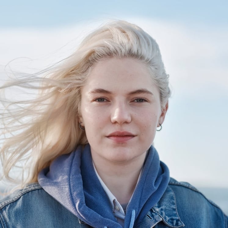 Woman with blond hair blowing in the wind, looking into the camera, with the sea behind her.